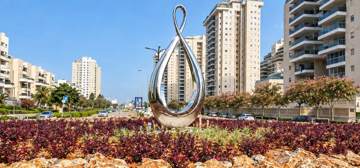 פסל נירוסטה מלוטש בצורת לולאת אינסוף גבוהה, בכיכר מול מגדלי מגורים באשדוד