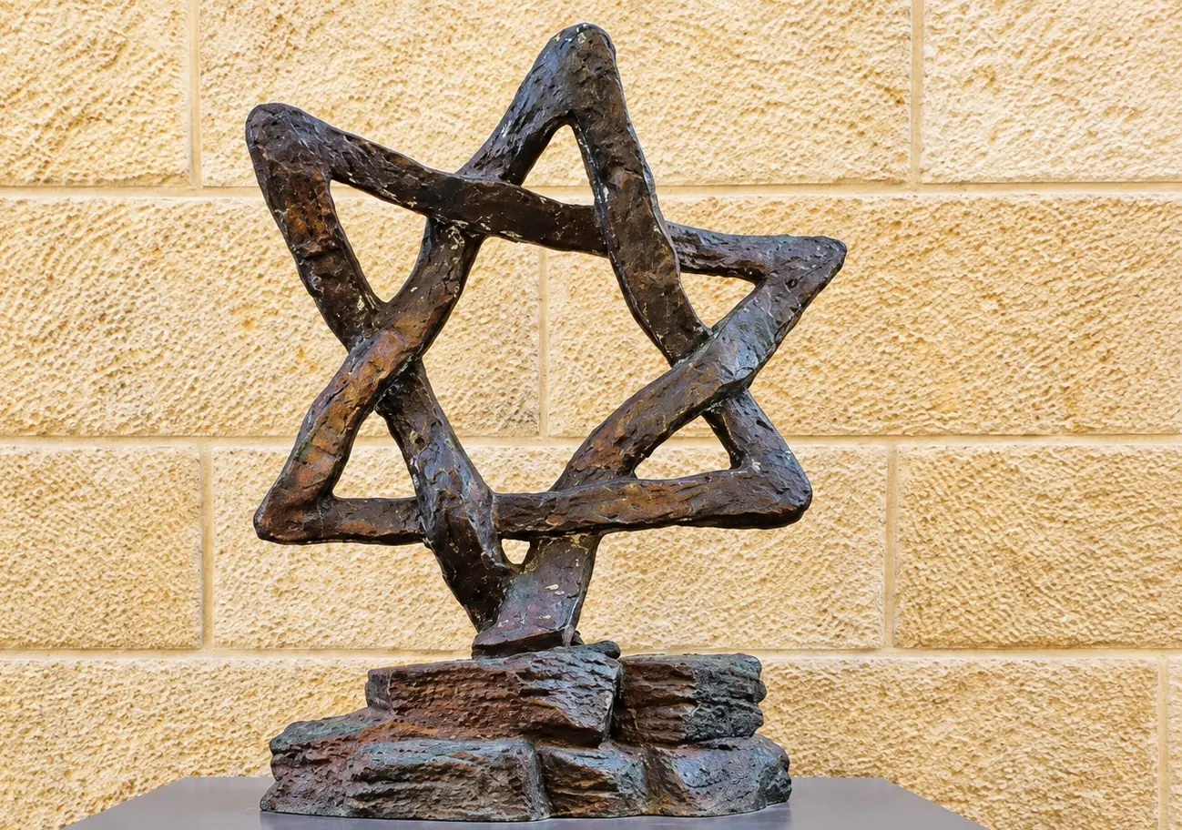 מגן דוד מיציקת ברונזה בעל רצועות מחוספסות, על ערימת סלעים מברונזה, מול קיר אבן ירושלמית